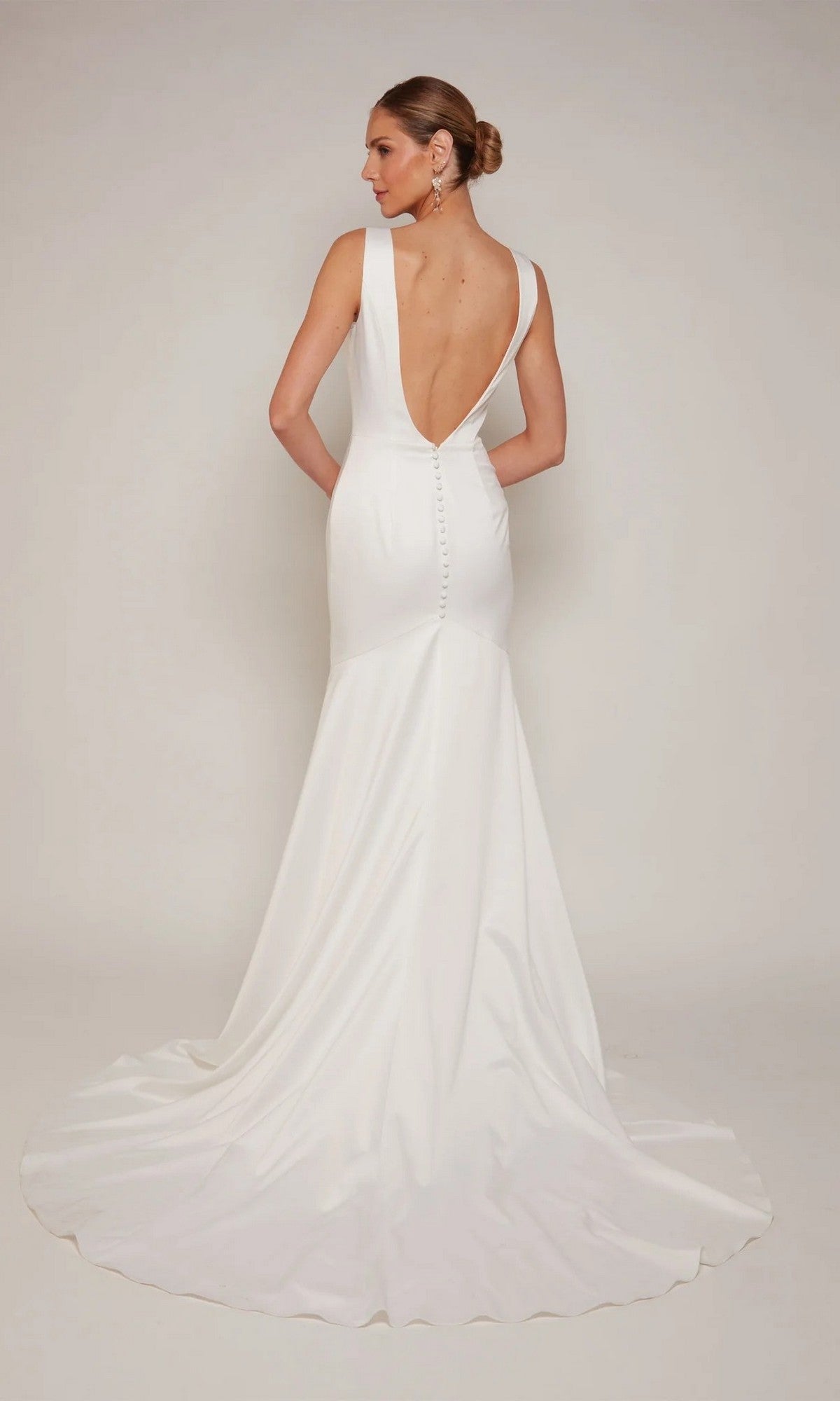 A woman in the Alyce 7091 long white dress—sleeveless with a plunging V-neckline, deep V-back, button accents, and a long train—stands against a plain light background.